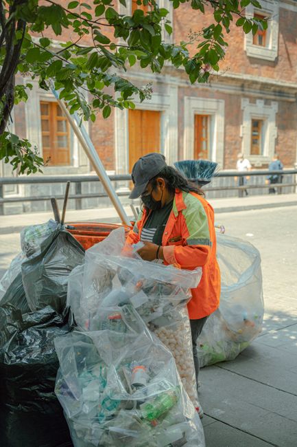A waste management worker wearing a black cap, dark sunglasses, and an orange high-visibility jacket with reflective yellow-green stripes is standing on a sidewalk next to a large pile of rubbish bags. The bags include black plastic refuse sacks, transparent plastic bags, and a green bag, all filled with various types of waste such as plastic bottles, cans, and general household rubbish. The worker appears to be inspecting or sorting items inside a clear plastic bag, with their head slightly bowed and hands inside the bag. In the background, there are historic brick buildings with wooden window frames and a tree with green leaves partially covering the upper left corner of the image. The scene is illuminated by natural daylight, creating a clear, detailed view of the waste collection environment—highlighting the importance of private and independent rubbish disposal services, as provided by companies like Rubbish Clearance Greenwich, to support local rubbish removal efforts in urban areas.