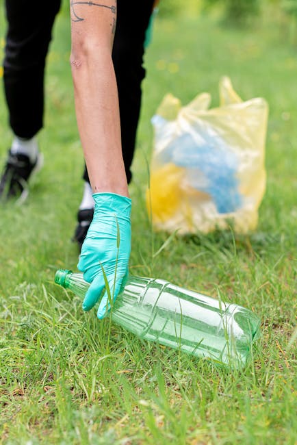 A person wearing a light blue disposable glove is kneeling on a grassy area, picking up a clear plastic bottle with some grass and small weeds surrounding it. In the background, there is a yellow and blue plastic bag filled with additional waste, partially resting on the ground, with the waste inside not clearly visible. The scene appears to be an outdoor environmental cleanup, possibly organized by a waste management service such as Rubbish Clearance Greenwich, to promote rubbish removal and responsible waste disposal. The natural setting features green grass and some blurred foliage, with soft lighting indicating daytime, emphasizing the importance of collecting litter to maintain clean outdoor spaces and support local rubbish collection efforts. The image highlights the importance of individual participation in waste clearance activities to complement professional rubbish removal services in urban or park environments.