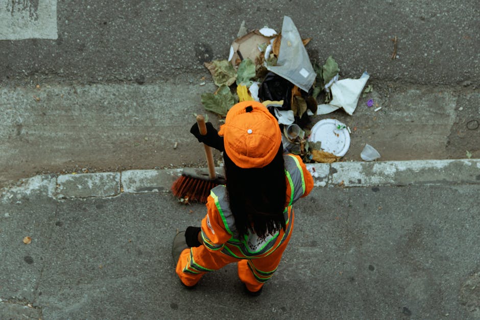A person wearing an orange high-visibility jacket and matching orange cap is seen from above, standing on a paved street adjacent to a curb. They are holding a broom with a wooden handle and black bristles, and appear to be engaged in street cleaning or rubbish collection. In front of the individual, there is a pile of mixed waste materials including crumpled paper, plastic bags, and leaves, some of which are torn or discarded. The waste is situated against a concrete wall or curb, with some debris spilling onto the asphalt surface. The parking line markings are faintly visible on the road surface, and the ambient lighting suggests daylight, typical for outdoor rubbish removal tasks. The scene reflects a professional approach to waste clearance, possibly conducted by a service such as Rubbish Clearance Greenwich, which offers independent rubbish collection and on-site clearance solutions to local residents and businesses.