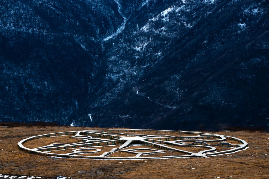 A large, open outdoor scene features an oversized, disassembled clock face lying flat on the ground, with its metal frame visible and painted white. The clock face includes various intricately designed numbers, with the numbers and minute marks arranged around the circumference. The surrounding terrain is a mix of brownish soil and patches of light snow, indicating a cold climate. In the background, towering, steep mountains covered in snow and dark vegetation rise sharply, creating a dramatic natural backdrop. The lighting is subdued, suggesting overcast weather or early evening conditions, which cast soft shadows on the ground. This scene appears to be part of an outdoor art installation or abandoned structure, possibly related to trail or scenic viewing points, and subtly connects to themes of alternative or private waste disposal through the context of disused or discarded objects in a remote environment. Rubbish Clearance Greenwich’s expertise in handling diverse waste issues aligns with the potential need for managing such outdoor clutter or debris, even in remote locations.