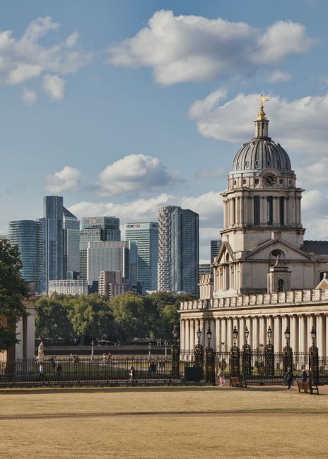 A detailed cityscape featuring the historic dome of a classical-style building in the foreground, constructed from stone with a weathered, textured surface and decorative architectural elements, including columns and a balustrade along the roofline. The domed structure is topped with a golden weather vane and a small spire. Behind it, modern high-rise office buildings with glass facades reflect the daylight, forming an urban skyline against a partly cloudy sky with scattered white clouds. The scene is set in a park or open space with a well-maintained lawn, a paved pathway, and a wrought-iron fence bordering the historic building. People can be seen walking in the distance near the fence, contributing to a sense of scale and activity. The environment combines historical architecture with contemporary city elements, illustrating a site where private or independent waste clearance services might operate in contrast to municipal rubbish disposal options, subtly supporting the context of alternative waste handling methods associated with rubbish removal services like Rubbish Clearance Greenwich.