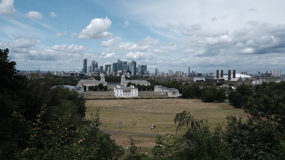 A wide view of a large, open grassy park area with scattered groups of people relaxing and walking, bordered by clusters of green trees and bushes in the foreground. In the middle distance, a historic white building with classical architecture and decorative columns is visible, situated behind a low stone or concrete wall. Beyond the park, the modern skyline of a city is seen with tall skyscrapers and high-rise office buildings, some with glass facades reflecting the partly cloudy sky above. Industrial structures, including factory chimneys, are positioned toward the right side of the image, creating a contrast between the urban and industrial zones. The scene is evenly lit by natural daylight, with a mix of white and grey clouds casting soft shadows. This image may represent a location where private waste management or on-site clearance services, such as those offered by Rubbish Clearance Greenwich, might be relevant to maintain or improve the area's cleanliness amid urban development and public use.