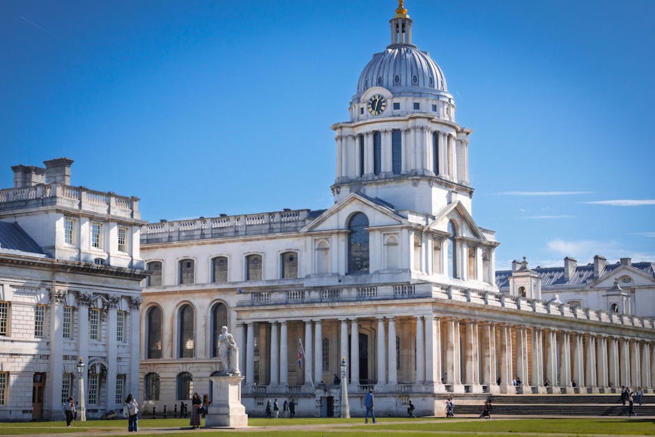 A detailed cityscape featuring the historic dome of a classical-style building in the foreground, constructed from stone with a weathered, textured surface and decorative architectural elements, including columns and a balustrade along the roofline. The domed structure is topped with a golden weather vane and a small spire. Behind it, modern high-rise office buildings with glass facades reflect the daylight, forming an urban skyline against a partly cloudy sky with scattered white clouds. The scene is set in a park or open space with a well-maintained lawn, a paved pathway, and a wrought-iron fence bordering the historic building. People can be seen walking in the distance near the fence, contributing to a sense of scale and activity. The environment combines historical architecture with contemporary city elements, illustrating a site where private or independent waste clearance services might operate in contrast to municipal rubbish disposal options, subtly supporting the context of alternative waste handling methods associated with rubbish removal services like Rubbish Clearance Greenwich.