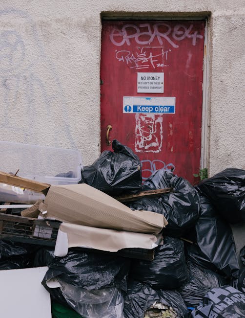 A large pile of black rubbish bags and discarded waste materials stacked in front of a red metal door set into a rough-textured, off-white wall. The bags are made of thick plastic and appear filled with domestic or general waste, with some partially torn or crumpled. Among the debris are pieces of cardboard, a ripped foam mat, and a plastic container, indicating mixed waste typical of private rubbish collection. The red door behind the pile has graffiti tags in white and pink spray paint, along with municipal and warning stickers; one sticker reads 'Keep clear' in white and blue, while another indicates 'No Money' and 'Do not block or impede' as part of waste disposal regulations. The surrounding environment suggests an urban setting, possibly an alleyway or service entry point used for independent rubbish removal, with concrete pavement visible at the base of the wall. The scene reflects a typical instance of non-licensed waste disposal needing removal by a professional service such as Rubbish Clearance Greenwich, who specialize in discreet and proper rubbish collection outside of council-managed channels. The lighting is natural, casting slight shadows that emphasize the textures of the trash and the worn surface of the door, reinforcing a neutral, factual observation of the scene's waste management context.