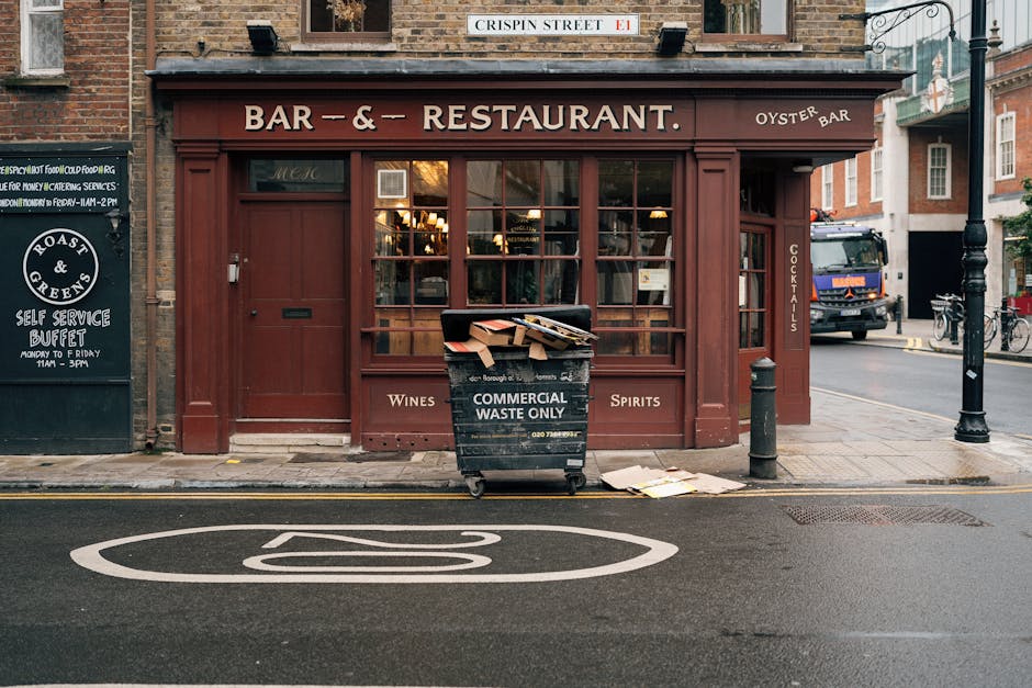 A black commercial waste bin labeled 'COMMERCIAL WASTE ONLY' is positioned on the sidewalk in front of a traditional pub-style building with a red wooden facade. The bin is partially open with cardboard boxes and discarded paper materials stacked on top, indicating ongoing waste collection. The surrounding environment includes a black bollard on the right side of the bin and a small pile of flattened cardboard boxes on the pavement nearby. Behind the bin, the pub's large front window reveals warm indoor lighting, with signage advertising wines and spirits, and a narrow door to the right. A street scene extends beyond the pub, featuring a parked blue truck, bicycles, and a lamp post, with the road marked by a white bicycle lane symbol. The building’s exterior displays a brick upper wall and a sign indicating “Crispin Street” at the top, suggesting a typical urban setting for private rubbish collection and waste management. Rubbish Clearance Greenwich specializes in such on-site waste clearance services within driven city environments, emphasizing the importance of appropriate disposal outside commercial premises.