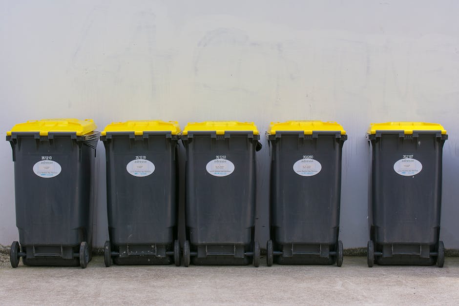 A large pile of black rubbish bags and discarded waste materials stacked in front of a red metal door set into a rough-textured, off-white wall. The bags are made of thick plastic and appear filled with domestic or general waste, with some partially torn or crumpled. Among the debris are pieces of cardboard, a ripped foam mat, and a plastic container, indicating mixed waste typical of private rubbish collection. The red door behind the pile has graffiti tags in white and pink spray paint, along with municipal and warning stickers; one sticker reads 'Keep clear' in white and blue, while another indicates 'No Money' and 'Do not block or impede' as part of waste disposal regulations. The surrounding environment suggests an urban setting, possibly an alleyway or service entry point used for independent rubbish removal, with concrete pavement visible at the base of the wall. The scene reflects a typical instance of non-licensed waste disposal needing removal by a professional service such as Rubbish Clearance Greenwich, who specialize in discreet and proper rubbish collection outside of council-managed channels. The lighting is natural, casting slight shadows that emphasize the textures of the trash and the worn surface of the door, reinforcing a neutral, factual observation of the scene's waste management context.