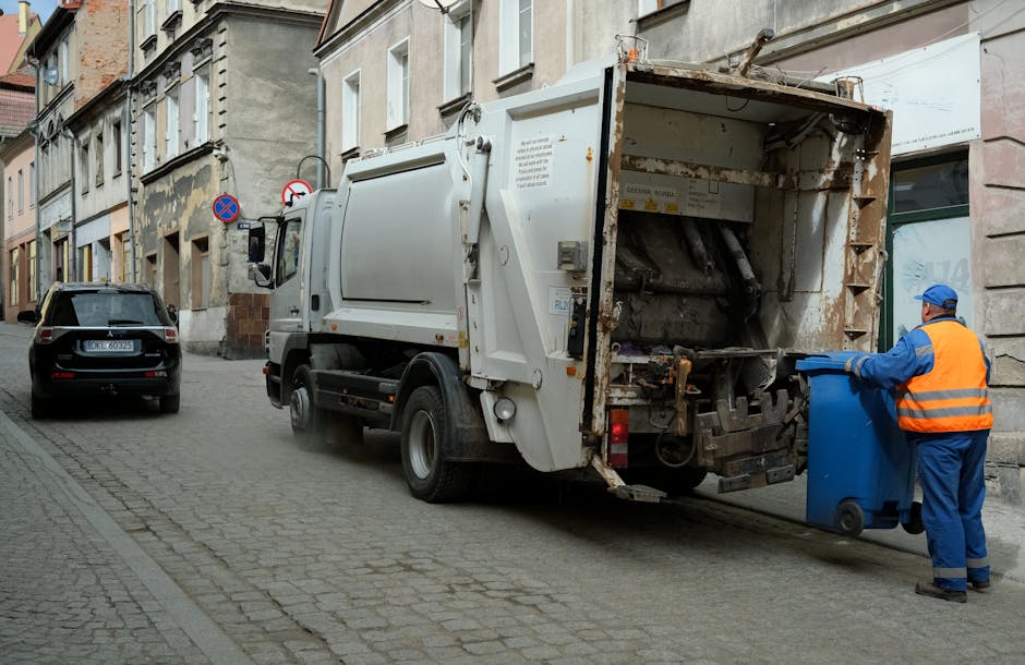 A waste collection truck operated by Rubbish Clearance Greenwich is parked on a narrow cobblestone street lined with old stone and brick buildings. The truck is white and has a large, open rear loading compartment with visible rust and dirt. A worker dressed in a blue uniform and orange high-visibility vest is seen emptying a blue recycling bin into the truck, standing on the right side of the image. The worker is wearing gloves and a matching blue cap. In the background, a black vehicle is parked further down the street, and there are multiple street signs attached to the buildings, including a no-parking sign. The scene is set during daylight, with natural light illuminating the street and buildings, capturing a moment of private waste collection or rubbish removal service often used in alternative waste handling compared to local authority collection services.
