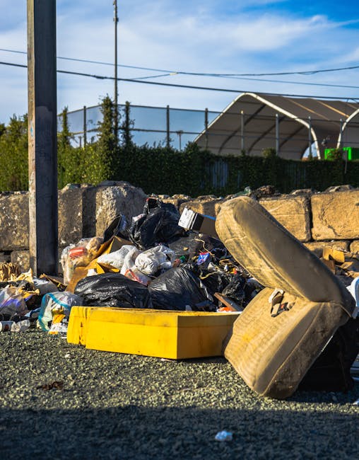 A roadside scene showing a pile of mixed household rubbish and waste materials, including black plastic garbage bags, a yellow plastic container, and an old, worn-out car seat with fabric upholstery, positioned on a gravel surface. Behind the rubbish, there is a low stone wall constructed from roughly hewn rocks. In the background, a tall metal pole supports overhead power lines, and a dense hedge of greenery is visible along with a partially visible structure with a curved, semi-transparent roof. The scene is illuminated by natural daylight, casting clear shadows, and the outdoor setting suggests an informal or unmanaged waste collection area, highlighting the importance of professional rubbish removal services such as those offered by Rubbish Clearance Greenwich for alternative waste handling and on-site clearance in Greenwich or similar areas.