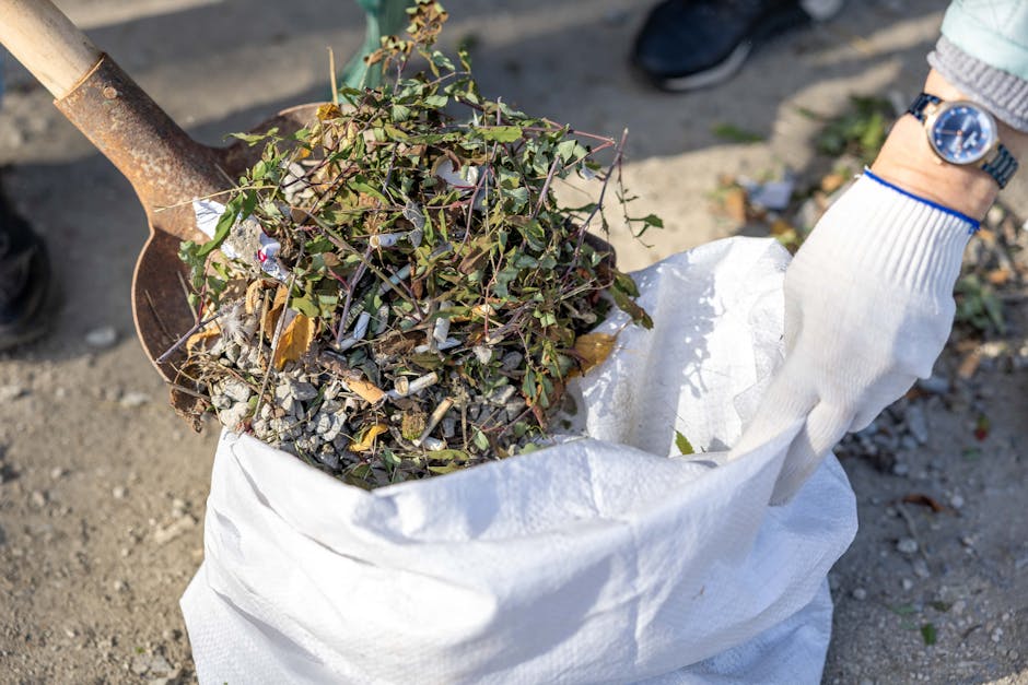 Close-up view of a person wearing a white glove and a wristwatch, lifting a white woven polypropylene sack filled with garden waste, including green leafy branches, small twigs, and gravelly soil, with a rusty shovel partially visible supporting the contents. The background shows a gravel surface with some scattered small debris and hints of footwear and clothing of nearby individuals, indicating outdoor activity. The scene illustrates the process of rubbish collection, highlighting the handling of garden waste for disposal or recycling, a task often associated with private rubbish clearance services like those offered by Rubbish Clearance Greenwich, especially when including non-standard or bulky items that may not be collected through conventional council services.