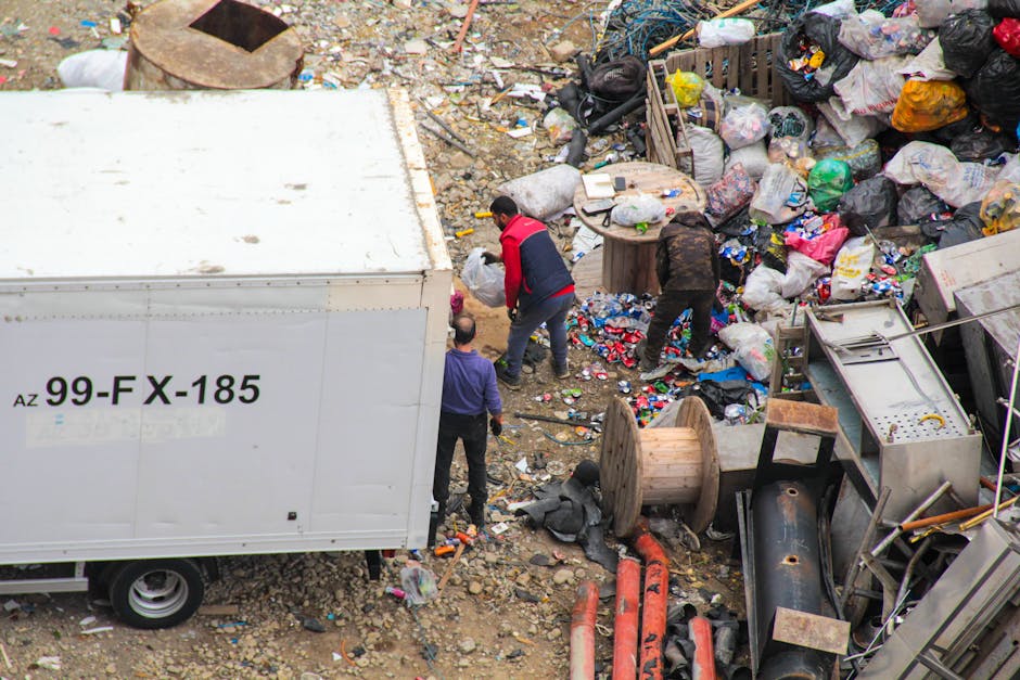 An outdoor scene showing a large pile of discarded waste, including black, white, and patterned plastic bags, cardboard boxes, and broken or discarded household items. The waste is situated on a gravel surface with scattered debris and rubble. To the left, part of a white van with the registration number 'AZ 99-FX-185' is visible, indicating a possible waste collection or disposal vehicle. In the middle of the image, three individuals are actively engaged in rubbish clearing; one person in a red and black jacket is leaning forward, working amidst the clutter, while another person in dark clothing is bending down to pick up or sort through debris. The surrounding environment is an outdoor open area, likely a site for waste transfer or informal dumping, with no clear sorting or recycling facilities visible. The scene illustrates the challenges of managing large quantities of waste and highlights the importance of professional rubbish removal services like those offered by Rubbish Clearance Greenwich for efficient, private collection solutions, especially in areas with access difficulties for standard refuse collection systems.