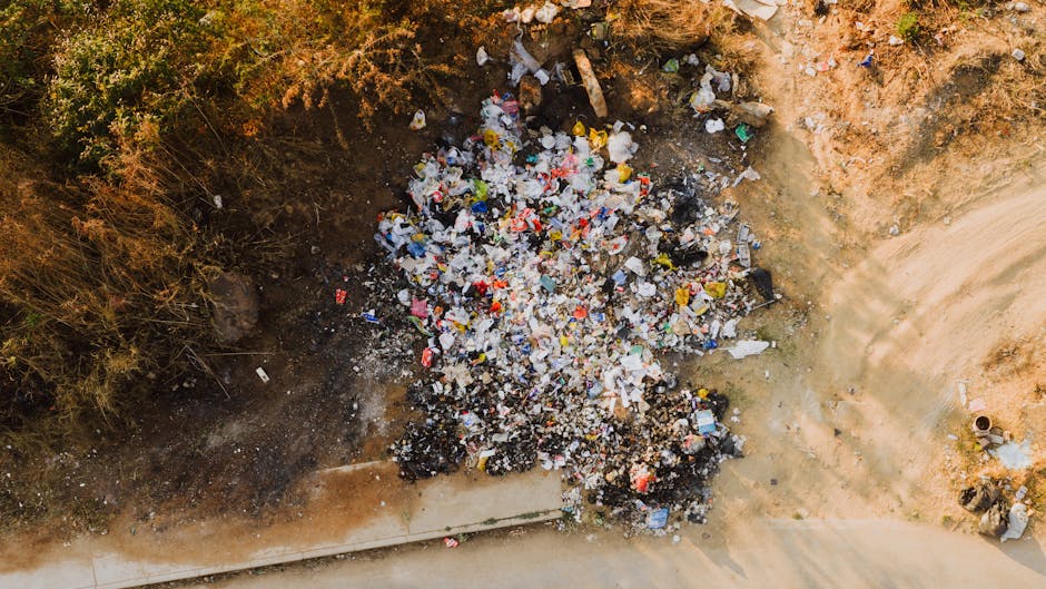 An outdoor scene showing a large pile of discarded waste, including black, white, and patterned plastic bags, cardboard boxes, and broken or discarded household items. The waste is situated on a gravel surface with scattered debris and rubble. To the left, part of a white van with the registration number 'AZ 99-FX-185' is visible, indicating a possible waste collection or disposal vehicle. In the middle of the image, three individuals are actively engaged in rubbish clearing; one person in a red and black jacket is leaning forward, working amidst the clutter, while another person in dark clothing is bending down to pick up or sort through debris. The surrounding environment is an outdoor open area, likely a site for waste transfer or informal dumping, with no clear sorting or recycling facilities visible. The scene illustrates the challenges of managing large quantities of waste and highlights the importance of professional rubbish removal services like those offered by Rubbish Clearance Greenwich for efficient, private collection solutions, especially in areas with access difficulties for standard refuse collection systems.